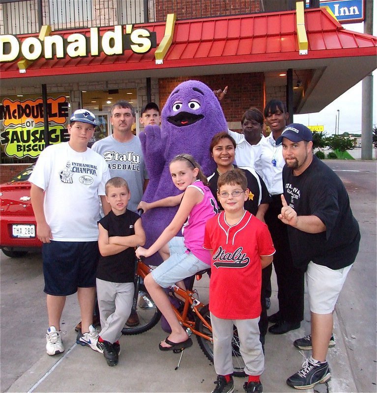 Image: IYAA players and coaches celebrate with McDonald’s® staff — Kirby Nelson wins the bicycle raffle during IYAA Sports Night at McDonald’s in Italy. Back row: IYAA president Gary Wood, Justin Wood, Grimace™ (Keanan Brown), Brenda Smith and Jakiesha Williams. Middle row: John Byers, Bryce DeBorde, Bike raffle winner Kirby Nelson, Maria Balderas and IYAA vice president Barry Byers. Front: Gage Wafer.