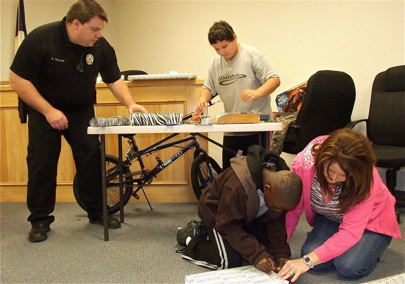 Image: Officer Tolliver helps Austin put the final touches on a wrapped present while Deonna Padilla assists Byron with the wrapping of one of his gifts.