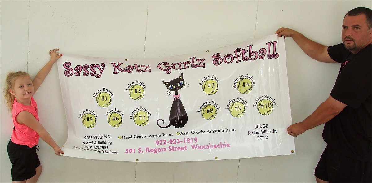 Image: Azlin Itson and her dad and coach, Aaron Itson, display the Sassy Katz Gurlz Softball team banner which shows the players and the team sponsors: Sassy Katz of Waxahachie, Cate Welding of Italy, Judge Jackie Miller, Jr. Precent 2. Lil’ Dab Embroidery of Italy added the numbers to the back of the team’s playoff uniforms.