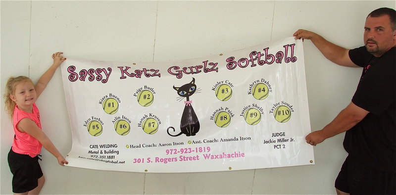 Image: Azlin Itson and her dad and coach, Aaron Itson, display the Sassy Katz Gurlz Softball team banner which shows the players and the team sponsors: Sassy Katz of Waxahachie, Cate Welding of Italy, Judge Jackie Miller, Jr. Precent 2. Lil’ Dab Embroidery of Italy added the numbers to the back of the team’s playoff uniforms.
