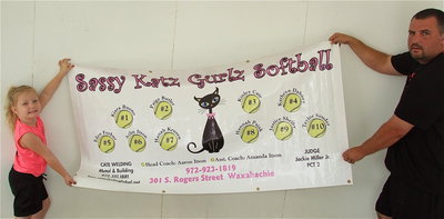 Image: Azlin Itson and her dad and coach, Aaron Itson, display the Sassy Katz Gurlz Softball team banner which shows the players and the team sponsors: Sassy Katz of Waxahachie, Cate Welding of Italy, Judge Jackie Miller, Jr. Precent 2. Lil’ Dab Embroidery of Italy added the numbers to the back of the team’s playoff uniforms.