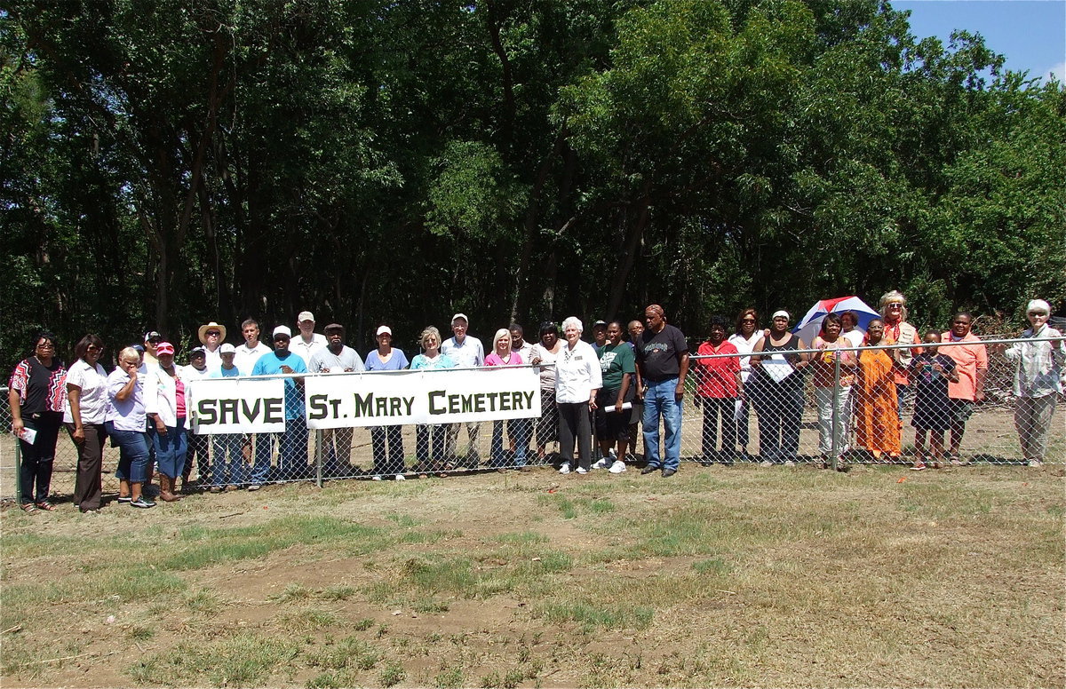 Image: Activists and guests who attended the SAVE St. Mary Cemetery rally in Ellis County, Texas pose around a sign hung by the Elmerine Bell Family and supporters on the fence that was added by Creek Land &amp; Cattle Company after the land developer destroyed a preexisting fence line while creating a road thru a portion of St. Mary Cemetery, destroying gravestones in the process that dated back to the 1870s.