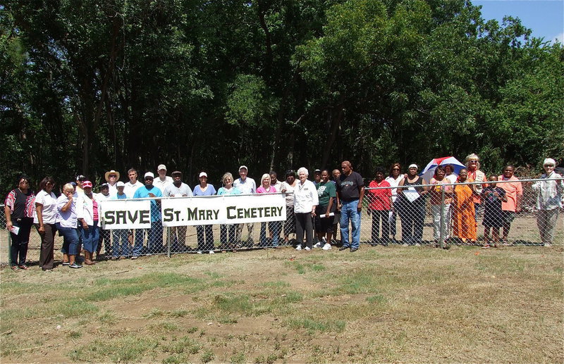 Image: Activists and guests who attended the SAVE St. Mary Cemetery rally in Ellis County, Texas pose around a sign hung by the Elmerine Bell Family and supporters on the fence that was added by Creek Land & Cattle Company after the land developer destroyed a preexisting fence line while creating a road thru a portion of St. Mary Cemetery, destroying gravestones in the process that dated back to the 1870s.