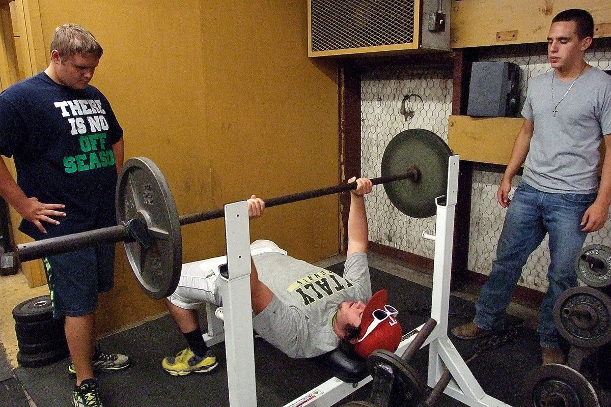 Image: Kyle Fortenberry does a practice rep on the bench as teammates Colin Newman and Cody Medrano ensure his safety.